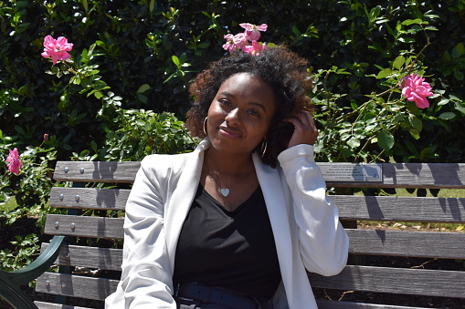 April 22, 2023, Hermann Park, Houston, Texas - Businesswoman of color wearing a white blazer sitting on a park bench with roses in the background.