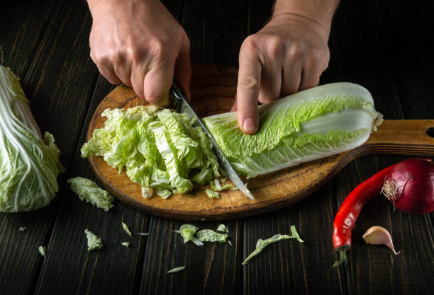istock Chef cuts fresh napa cabbage with knife for salad on a vintage kitchen board with fresh vegetables. Cooking and restaurant or cafe concept 1484307955