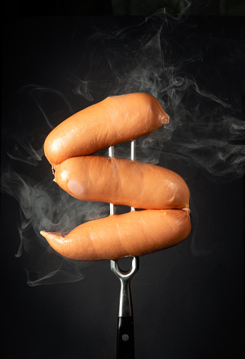 Thick boiled milk sausages with steam on a fork on a black background. Street hot food concept.