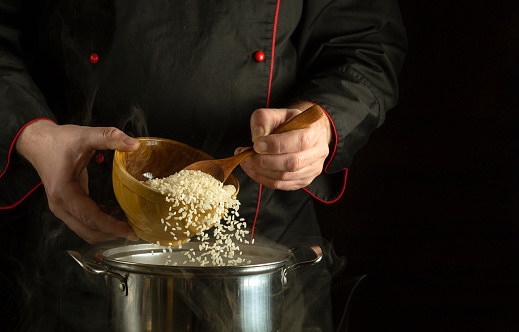 Professional chef spoons the groats of rice into the pan. Dark space for recipe or menu. The idea of cooking delicious rice porridge in a restaurant kitchen