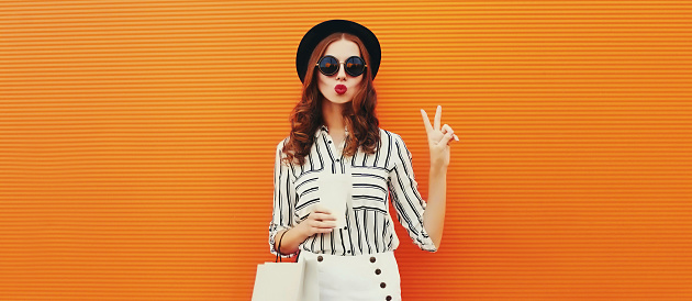 Portrait of beautiful young woman model with shopping bags and cup of coffee blowing her lips sending air kiss wearing white striped shirt, black round hat on orange background