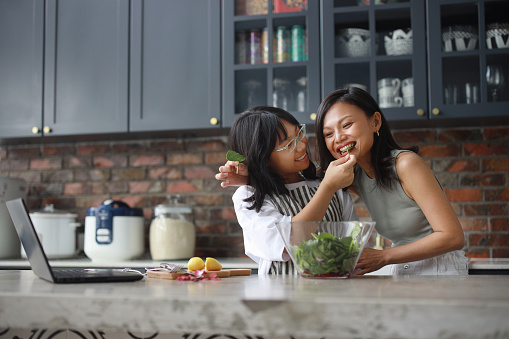 A cheerful Asian mother and daughter are using a laptop computer in the kitchen and having fun while mixing salad in a bowl