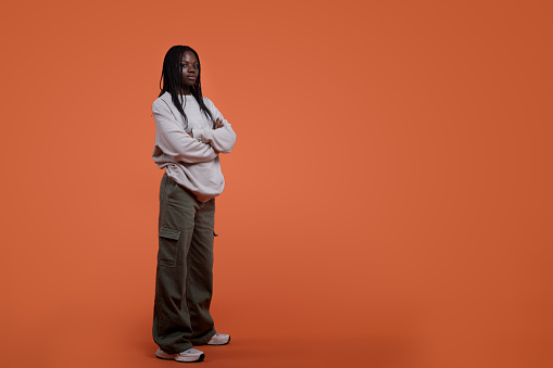 Full body of serious young African American female model in casual wear with crossed arms and braided hair standing against orange background and looking at camera in bright studio