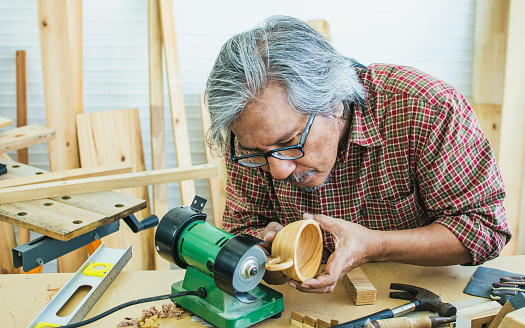 Senior Asian happy retired old male woodworker or carpenter smiling with happiness, wearing check shirt and eyeglasses, creating DIY wooden coffee cup for house decoration. Hobby, Retirement Concept