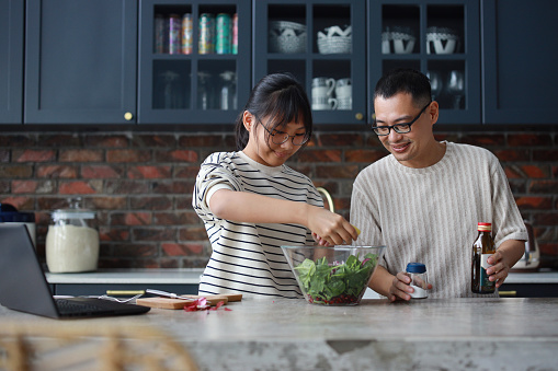 An Asian father and daughter are bonding in the kitchen on the weekend, as they prepare a salad together while using a laptop computer