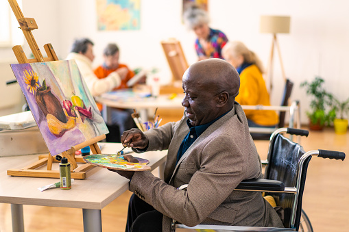 A retired man in a wheelchair is painting onto a canvas, enjoying an art class in a bright modern room, more people in the background are also painting