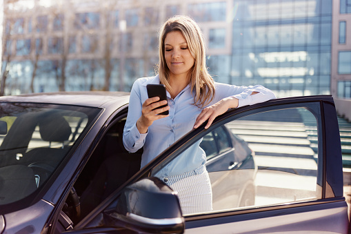 Young female entrepreneur reading a text message on smart phone while standing by her car on the street.