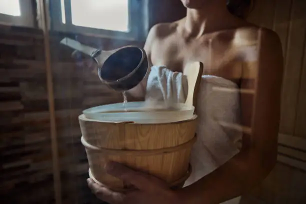 Close up of unrecognizable woman holding a water bucket in sauna. The view is through glass.