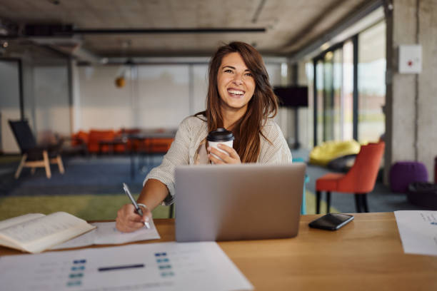 Happy creative woman taking notes while working on laptop in the office.