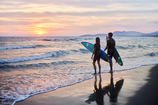 Rear view of couple of surfers with their surfboards during summer sunset on the beach. Copy space.