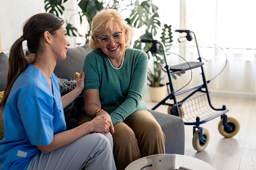 Female home care specialist and satisfied elderly woman holding hands while talking and sitting in living room. Happy senior woman enjoying company of her nurse during regular home visit.