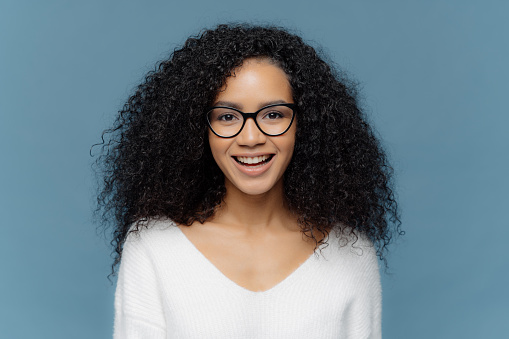 Photo of cheerful dark skinned female with bushy crisp hair, smiles happily at camera, rejoices nice day, dressed in white sweater, isolated over blue background expresses positiveness. Human feelings