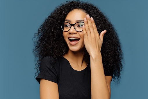 Headshot of beautiful curly woman looks aside, touches face, wears transparent glasses, black casual t shirt, isolated on blue background, notices something pleasant. People and positiveness concept