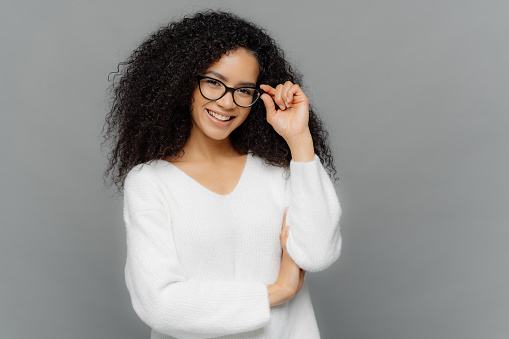 Portrait of happy woman with curly hair, kepes hands on frame of glasses, has gentle smile, wears white jumper, isolated over grey background. Copy space for your advert. Delighted student indoor