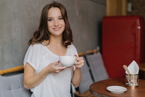 Smiling woman with tender smile, holds cup of tea or coffee, enjoys good rest, poses in cozy cafe or restaurant, waits for friend, dressed in casual outfit. People, rest and lifestyle concept