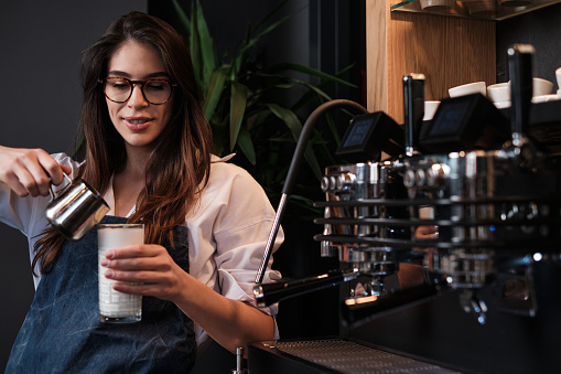 A happy barista preparing fresh coffee and adding milk into a glass while standing next to a coffee machine.