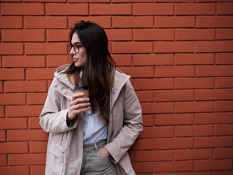 A happy smart casual woman leaning on brick wall outside and enjoying takeaway coffee.