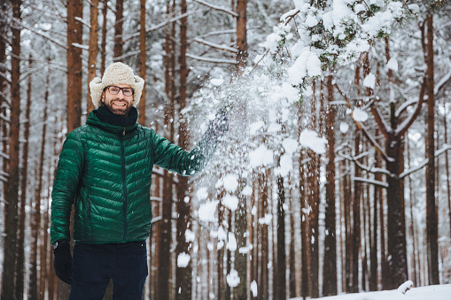 Smiling delightful male dressed in warm clothes, stands in winter forest, throws snow in air, has fun alone, has good mood, expresses positive emotions and feelings. Positiveness concept