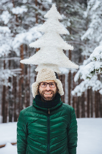Vertical portrait of cheerful bearded man has fun alone in winter forest, keeps artificial fir tree, poses outdoors, admires frosty snowy weather, expresses positiveness and pleasant emotions