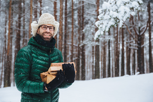 Cheerfu pleasant looking beard male model holds firewood, spends free time in forest, going to fry shish kebab, has picnic in family circle, logs wood, enjoys fresh winter forest air outdoor