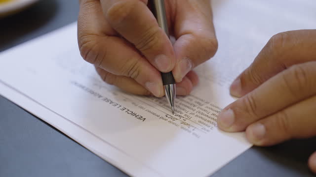 Close-up of a man's hand filling out personal information in a vehicle lease agreement using black ink pen and putting then putting the pen down.