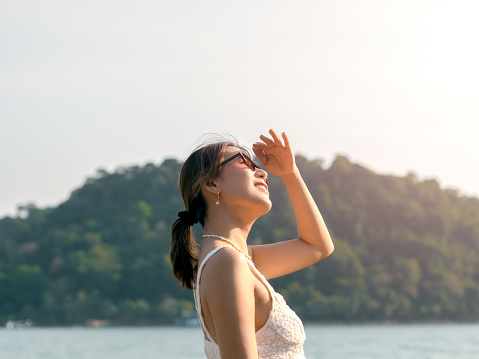 Beautiful Asian woman with sunglasses on the beach in summer vibes. Happy smile female covering the sunshine with her hand on the sea, green mountain island and sky background, Holiday vacation.