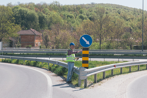 Road worker inspecting signs and road markings on highway intersection using digital tablet