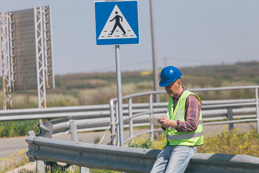 Road worker inspecting signs and road markings on highway intersection using digital tablet