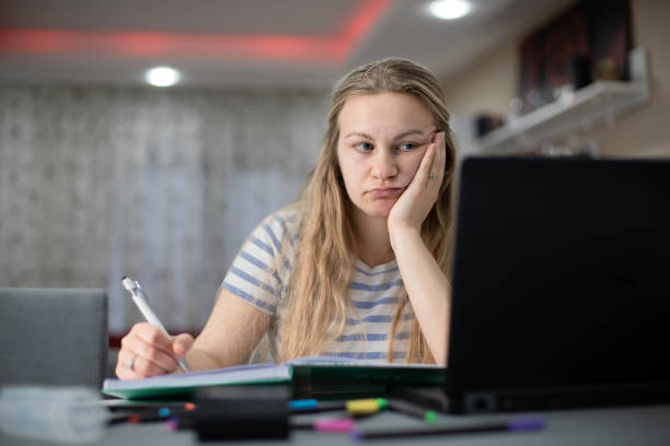 istock Young bored student woman sitting at desk and learning , she is using computer and writing in notebook 1484467131