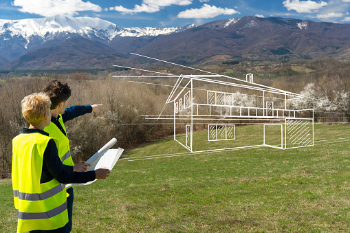 A young couple holding blueprints and wearing fluorescent vests are pointing into the distance as they imagine their new home on the hill with mountains in the background