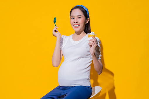 Happy pregnant asian female young woman enjoy eating ice cream studio shot on yellow background happy and smiling,beautiful young pregnant woman love her sweet desert eating an ice cream
