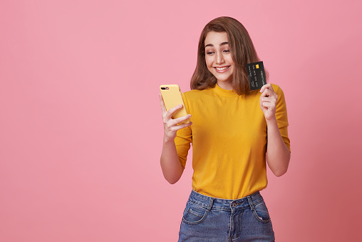 happy smiling young woman using mobile phone with credit card for shopping and payment online isolated on pink studio background.