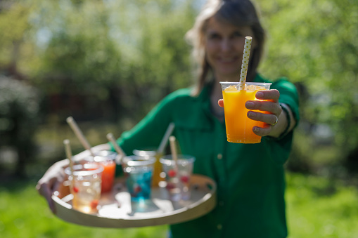 Woman carrying recyclable transparent PET (polyethylene terephthalate) cups with colorful juice and soft drinks on a wooden tray. The woman is handing a soft drink to the camera.
Photographed outdoors with shallow depth of focus.
Canon R5