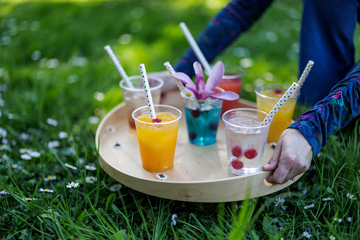 Woman carrying recyclable transparent PET (polyethylene terephthalate) cups with colorful juice and soft drinks on a wooden tray.\nPhotographed outdoors with shallow depth of focus.\nCanon R5