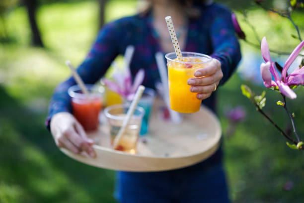 Woman carrying recyclable transparent PET (polyethylene terephthalate) cups with colorful juice and soft drinks on a wooden tray. The woman is handing a soft drink to the camera.Photographed outdoors with shallow depth of focus.Canon R5