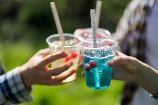 Three people holding recyclable transparent PET (polyethylene terephthalate) cups with colorful water, juice and soft drinks. They are making a toast.\nPhotographed outdoors with shallow depth of focus.\nCanon R5