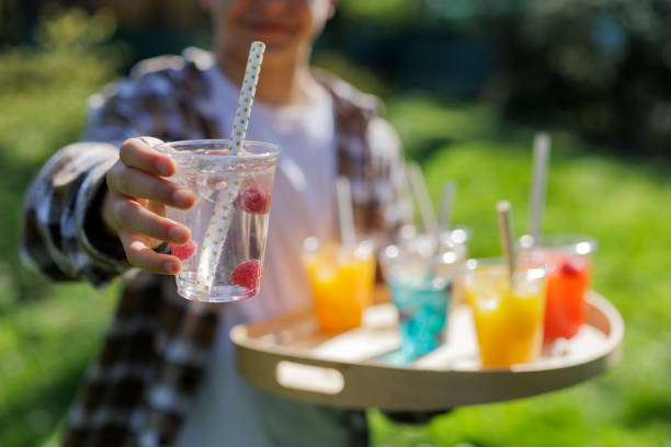 Teenager carrying recyclable transparent PET (polyethylene terephthalate) cups with colorful juice and soft drinks on a wooden tray. The teenager is handing a water drink to the camera.Photographed outdoors with shallow depth of focus.Canon R5