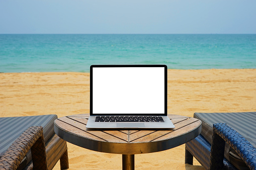 Laptop with blank screen for creative design on the table nearby sea and beach background. Computer notebook with monitor clipping path for present landing page design. Laptop computer mock up template