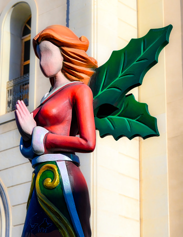 Alicante, Spain - January 5, 2023: Statue of a faceless woman with her hands clasped beneath her breasts. The statue is adorned with two green wing-like structures resembling leaves and is in front of a building.