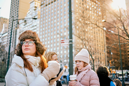Young woman in warm clothing holding takeaway coffee looking way while outside in the city on an autumn day. Girl having coffee with her mother and sister standing back in the city on a winter day.