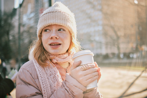 Portrait of a teenage girl in warm wear having coffee standing outside in the city. Small girl holding hot coffee outdoors on a cold winter day.
