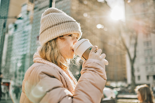 Teenage girl in warm wear drinking coffee standing outside in the city on winter day. Girl having hot coffee outdoors on a cold autumn day.
