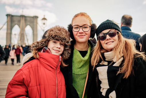 Portrait of a beautiful family of three winter wears standing together on Brooklyn bridge on a winter day. Woman with her two girls in warm clothing looking at camera and smiling in the city.