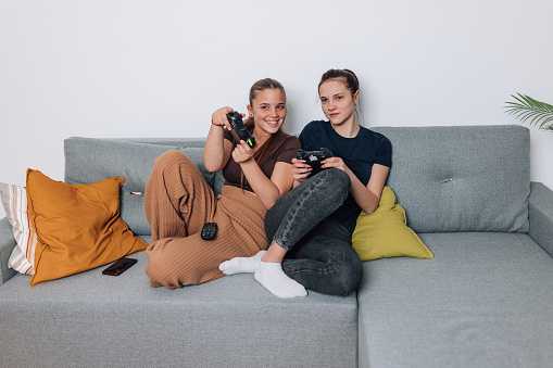 Two teenage sisters enjoy an afternoon of friendly competition as they play video games with joysticks while sitting on the sofa