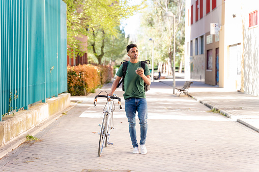 Full body side view of bearded male in casual clothes with backpack walking along street with bicycle and looking away