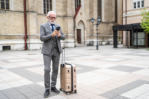 Business trip, senior businessman outside hotel using phone, mature boss with suitcase, investor in business suit and glasses