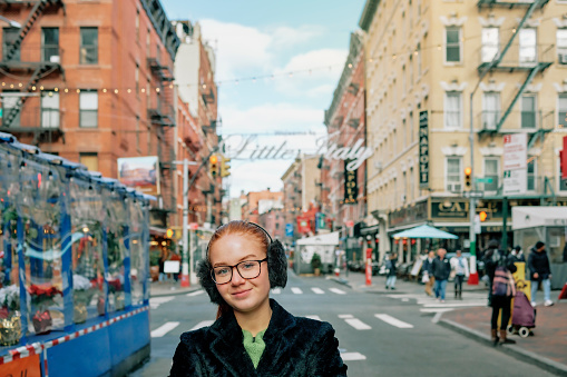 Portrait of a beautiful young girl in warm jacket and ear muffs standing along the city street. Young woman looking at camera and smiling standing in the city on an autumn day. Teenager in Little Italy, New York.