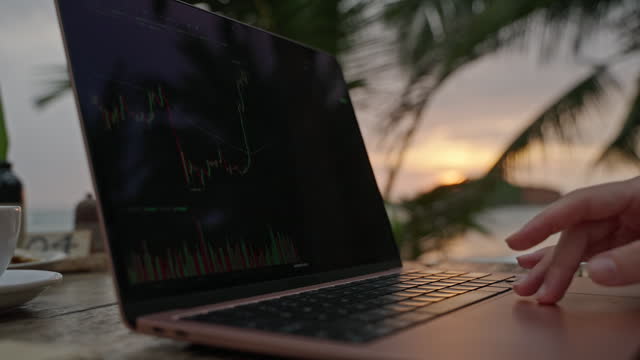 Female cryptocurrency trader at laptop checks charts online working remotely at outdoor tropical seaside cafe at sunset. Woman crypto broker analyses graphic of stock exchange rates by ocean close-up