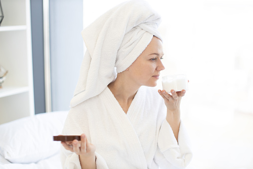 Close-up of lovely carefree lady in towel savouring pleasant fragrance of cosmetic product in hand while pampering herself with skincare procedures. Facial cream moisturizing skin after showering.
