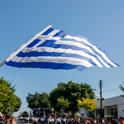 Greek flag fluttering in front of blue sky over crowded street during parade